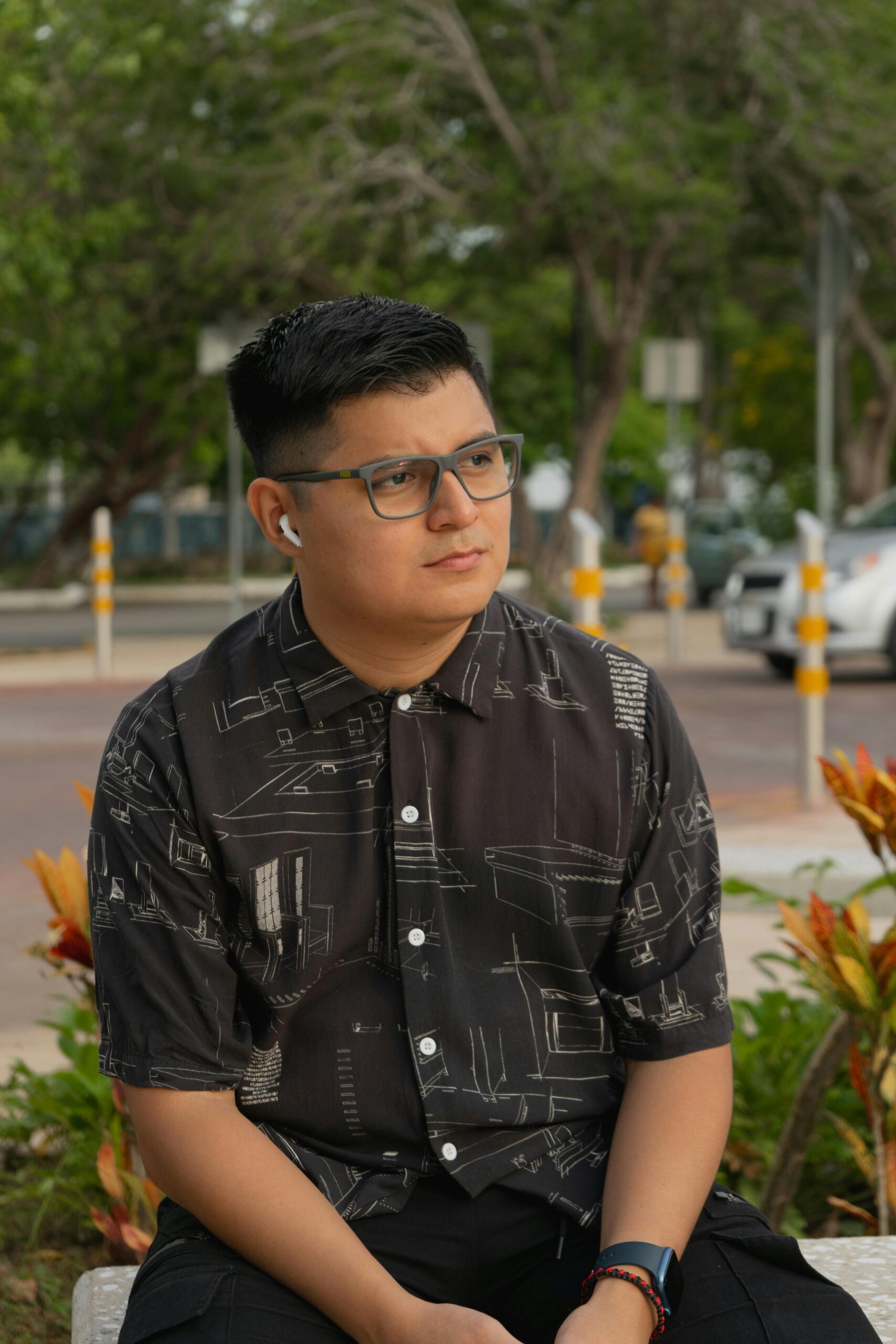 A man with glasses sits in a park wearing a patterned shirt and using earbuds.